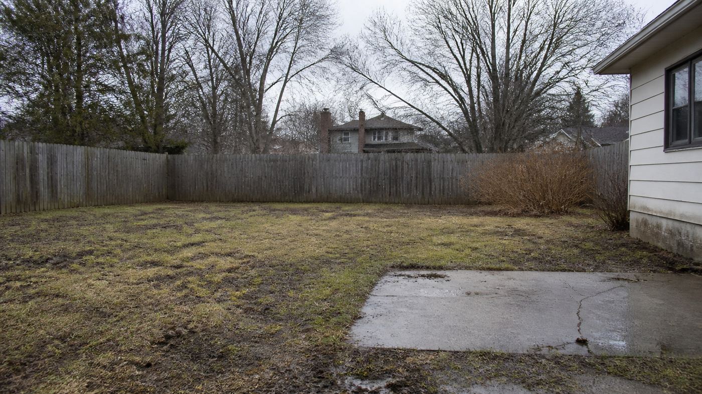 Plain neglected muddy backyard with patchy grass and cracked concrete slab, overcast day, BEFORE renovation shot, same camera angle as after